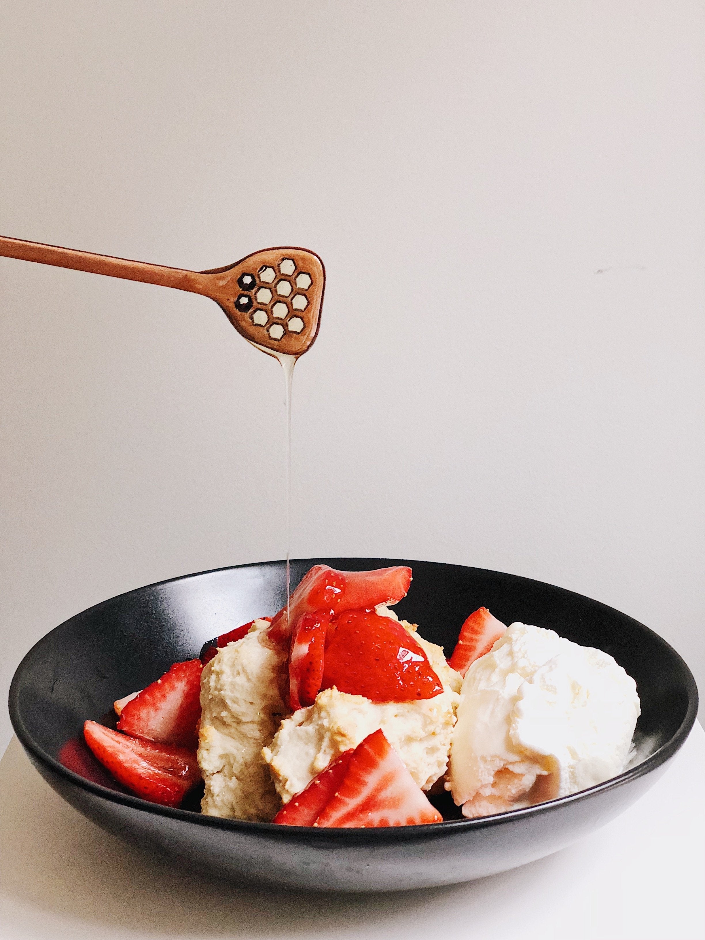 wooden honey dipper dripping honey over a black shallow bowl of shortcake with strawberries and cream