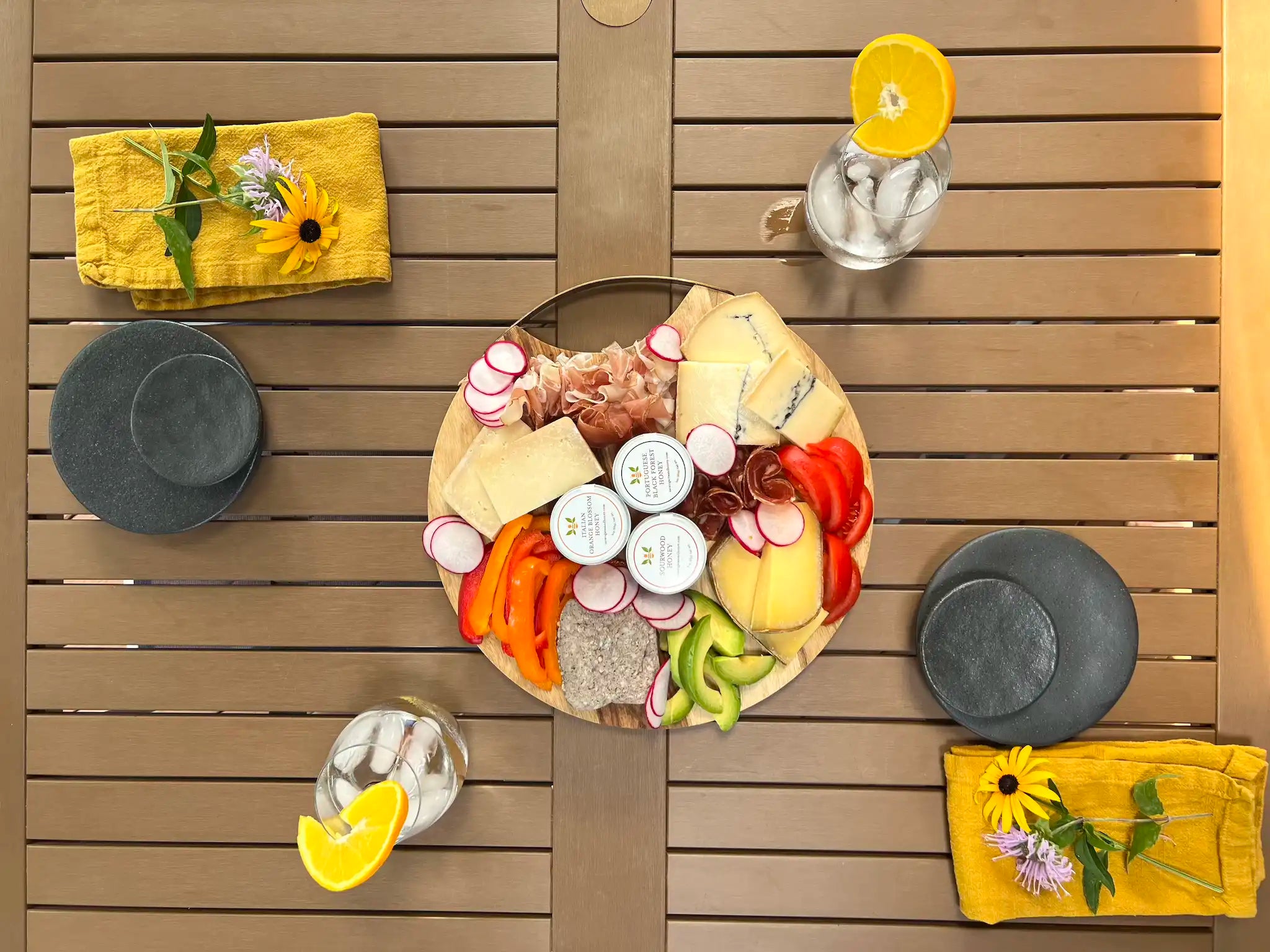 aerial view of a charcuterie and cheese board with three mini honeys between two place settings with yellow napkins on an outdoor table