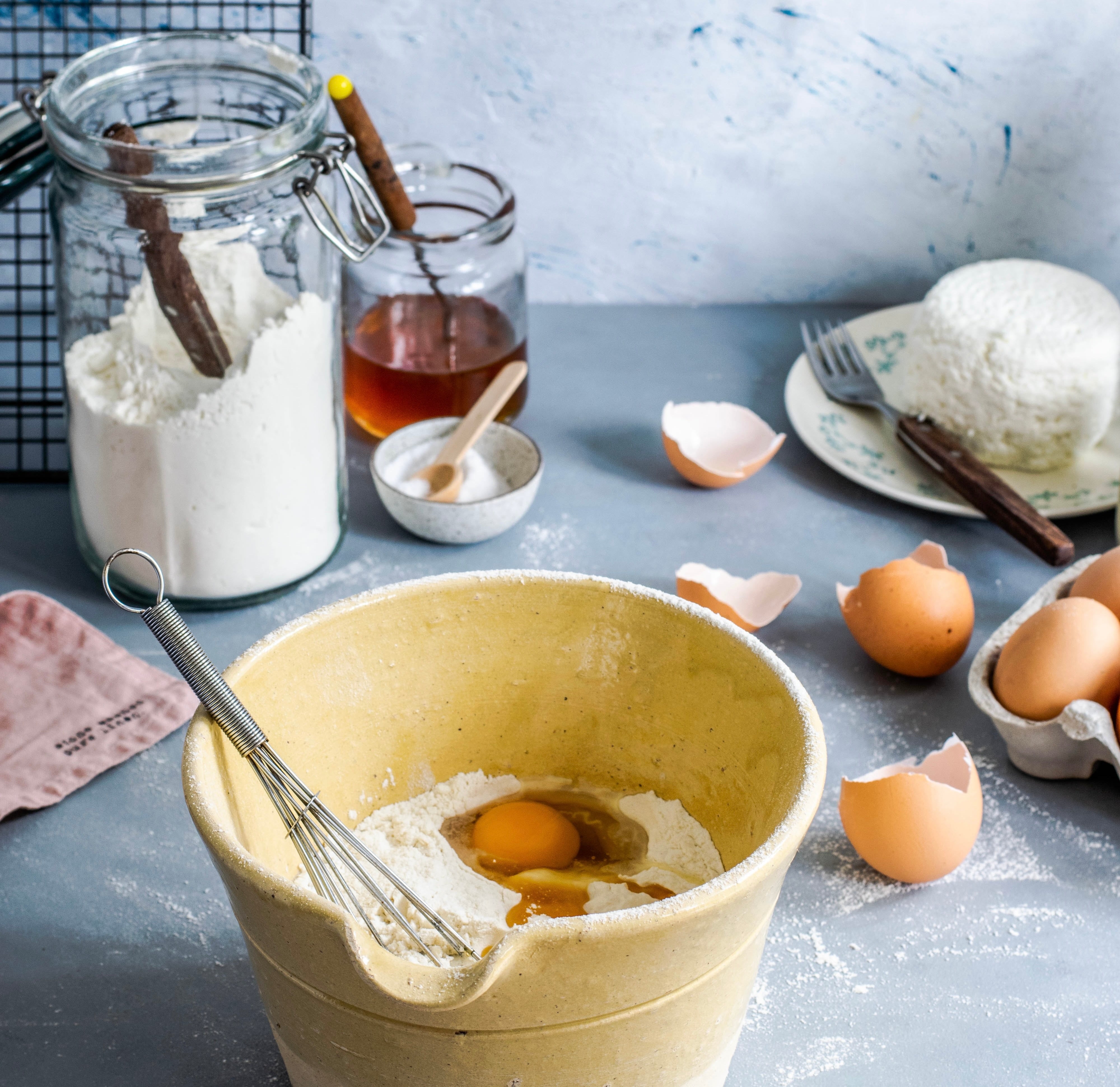 artistically messy baking counter with a yellow ceramic mixing bowl in the foreground filled with flower, eggs, and a whisk. Honey, salt, butter and eggs are arrayed in the background