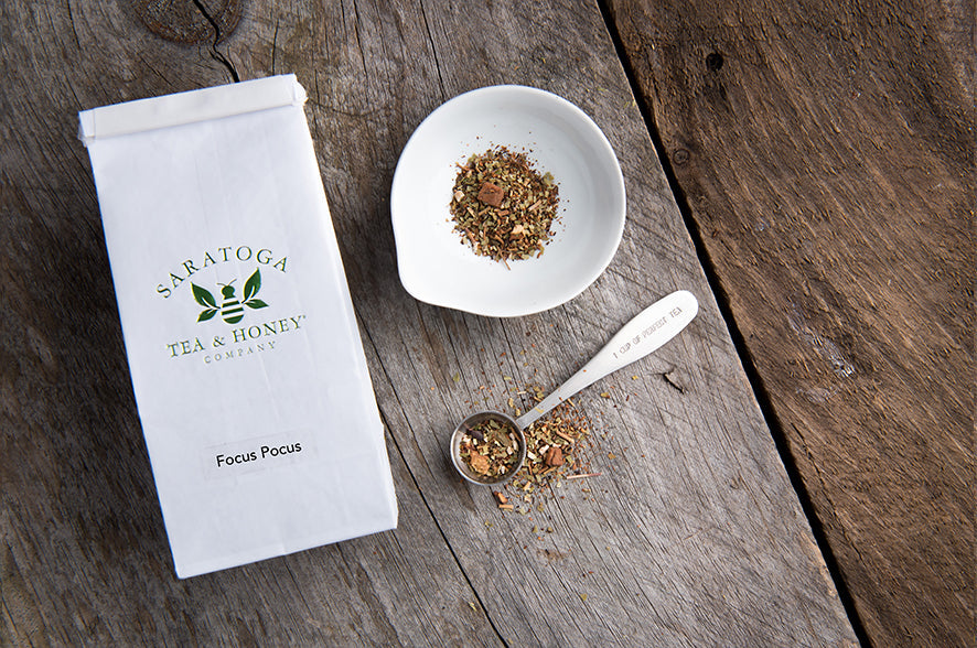 white tin tie bag of Focus Pocus next to a small saucer of loose leaf tea and a metal scoop spilling tea on a wooden background