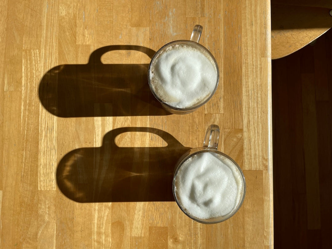 two staggered mugs of london fog lattes topped with milk foam. The sun is coming from the right and casting stark shadows from the mugs onto the wood background