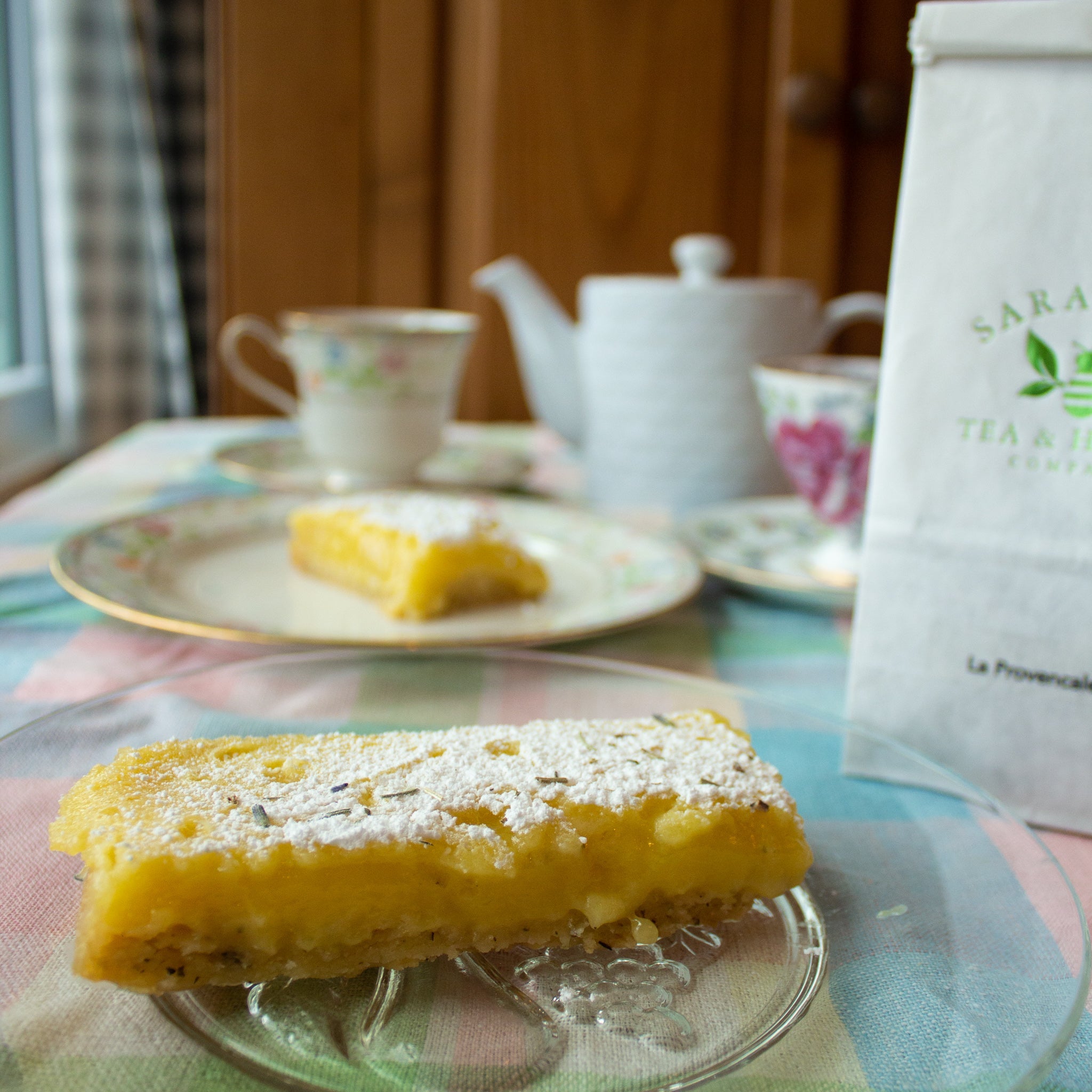 gooey, bright yellow lemon bar on a glass plate in front of a bag of La Provençale, another plate of lemon bars, and a vintage tea set