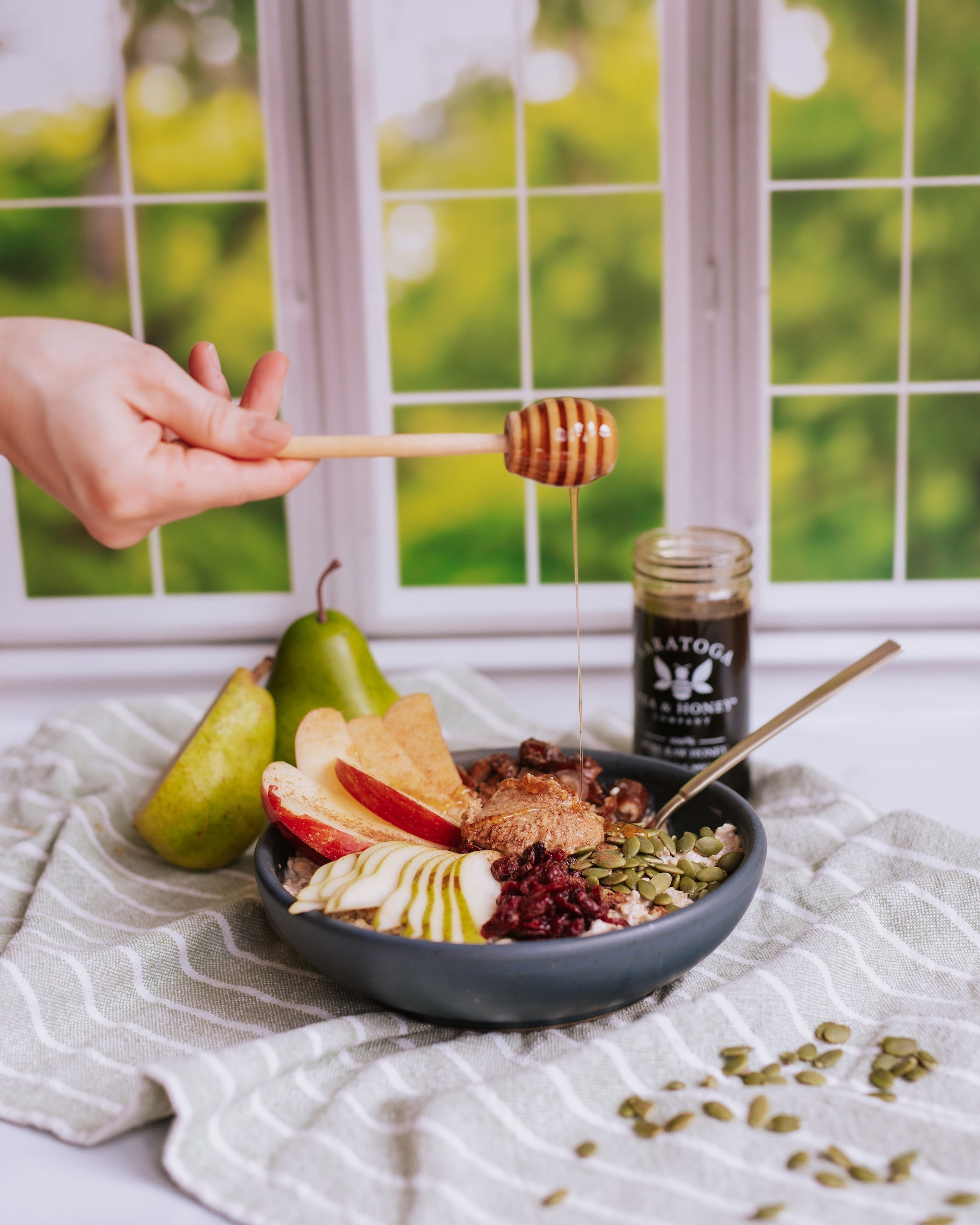 hand drizzling honey over an oatmeal power bowl with sliced pear, sliced apple, almond butter, dried cranberies, and pumpkin seeds