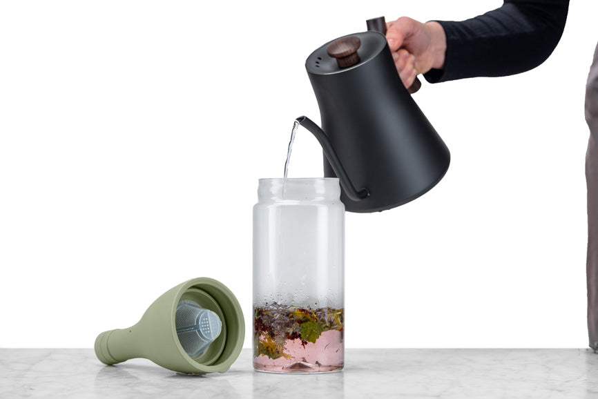 hand pouring water from a black gooseneck kettle into a glass bottle for brewing tea