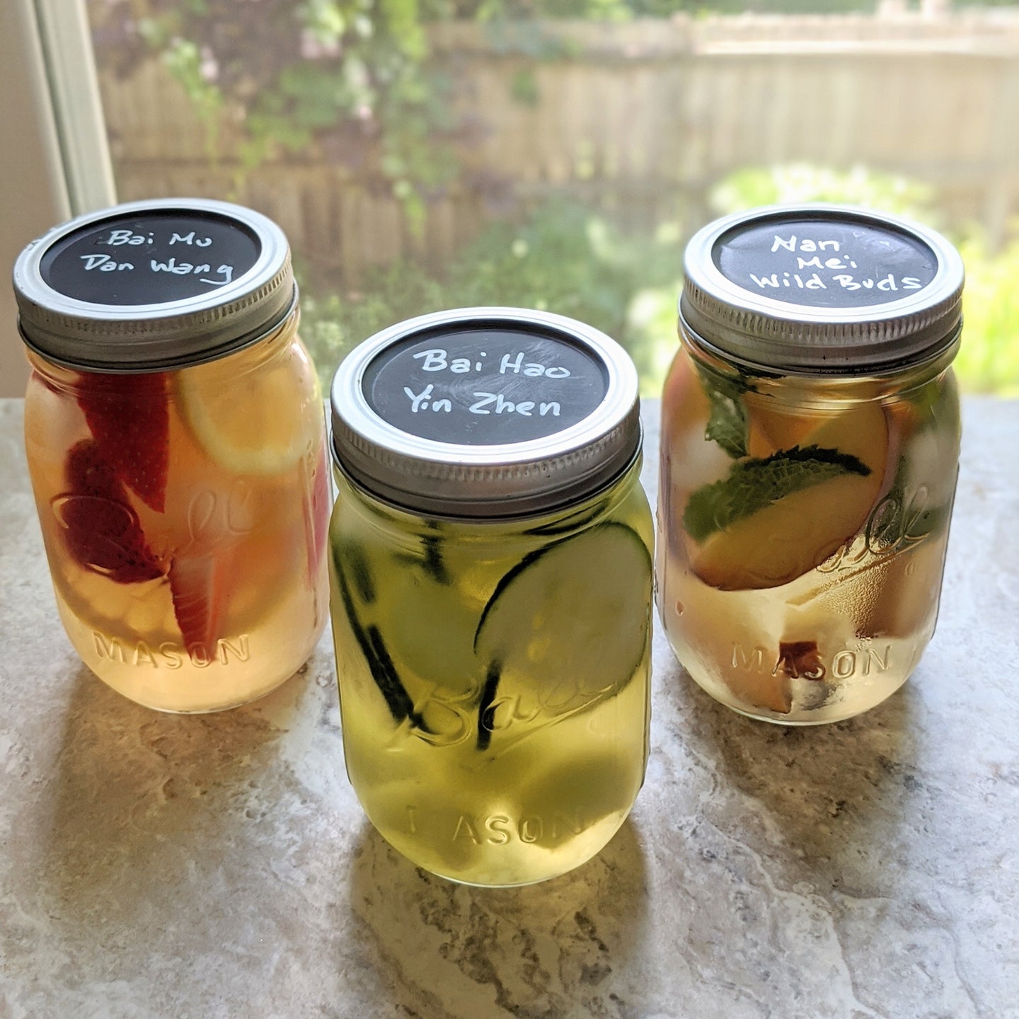 three mason jars filled with fruit infused tea; from left to right: bai mu dan with strawberries and lemon, bai hao yin zhen with cucumber, and nan mei wild buds with peaches