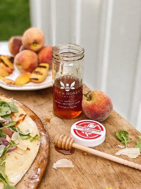 jar of Saratoga Tea & Honey Co. Ghost Pepper Honey on a wooden table next to a pizza with grilled peaches in the background