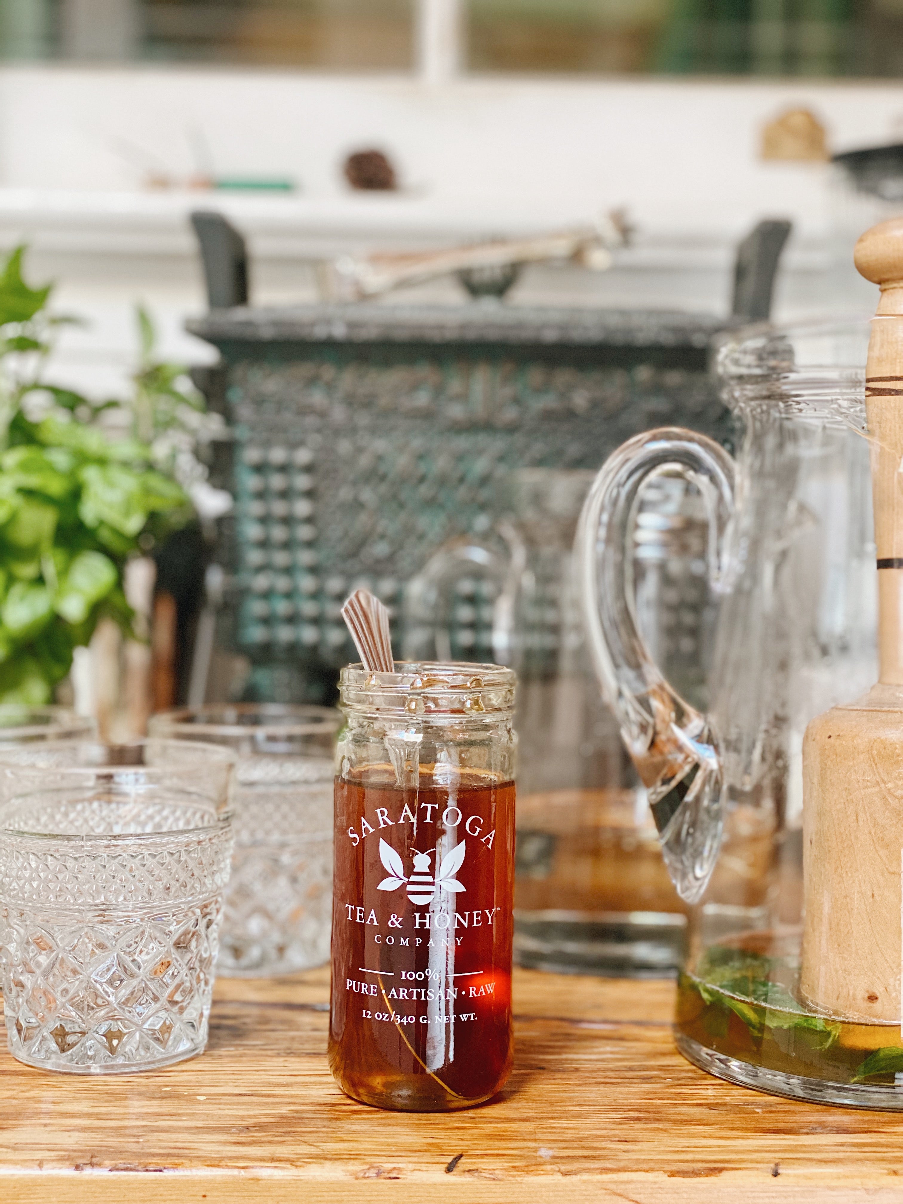 jar of Saratoga Tea & Honey Co Ghost Pepper honey with a spoon set amidst lowball glasses and a glass pitcher on a butcher block counter