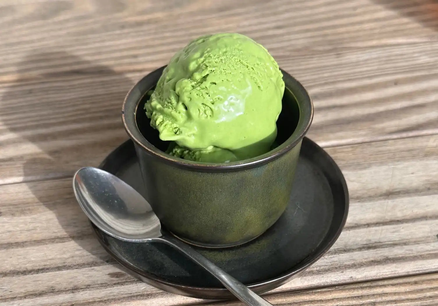 scoop of bright green, creamy matcha ice cream in a black cup with matching saucer and sliver spoon on a picnic table background