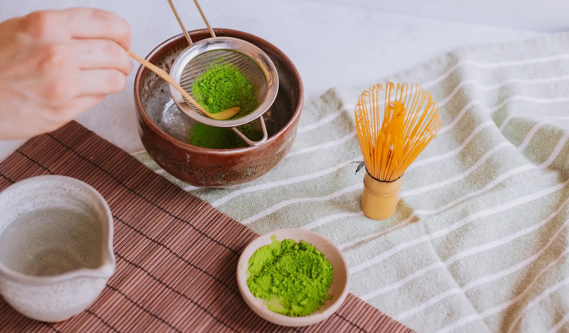 https://www.saratogateaandhoney.com/cdn/shop/files/Matcha_teaware_with_hand_sifting_matcha_into_a_chawan_with_a_chashaku_and_chasen_in_the_foreground.webp?v=1680788371&width=3840