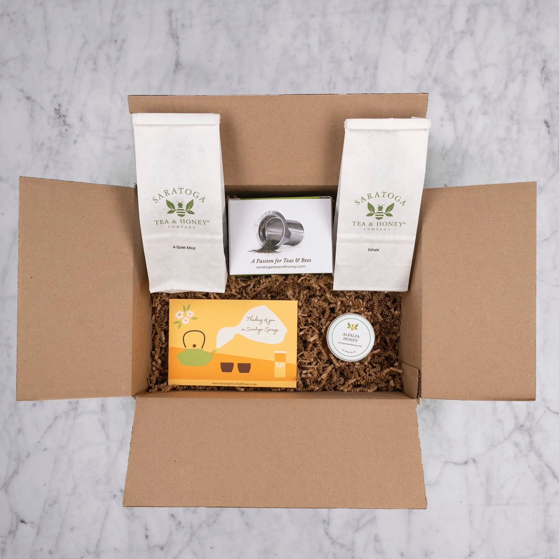 Showing contents of tea and honey sympathy gift in a brow box with gift fill. Includes: 2oz bag of A Quiet Mind, 2oz bag of Exhale, stainless steel tea infuser, Alfalfa mini honey, and a thinking of you tea and honey post card