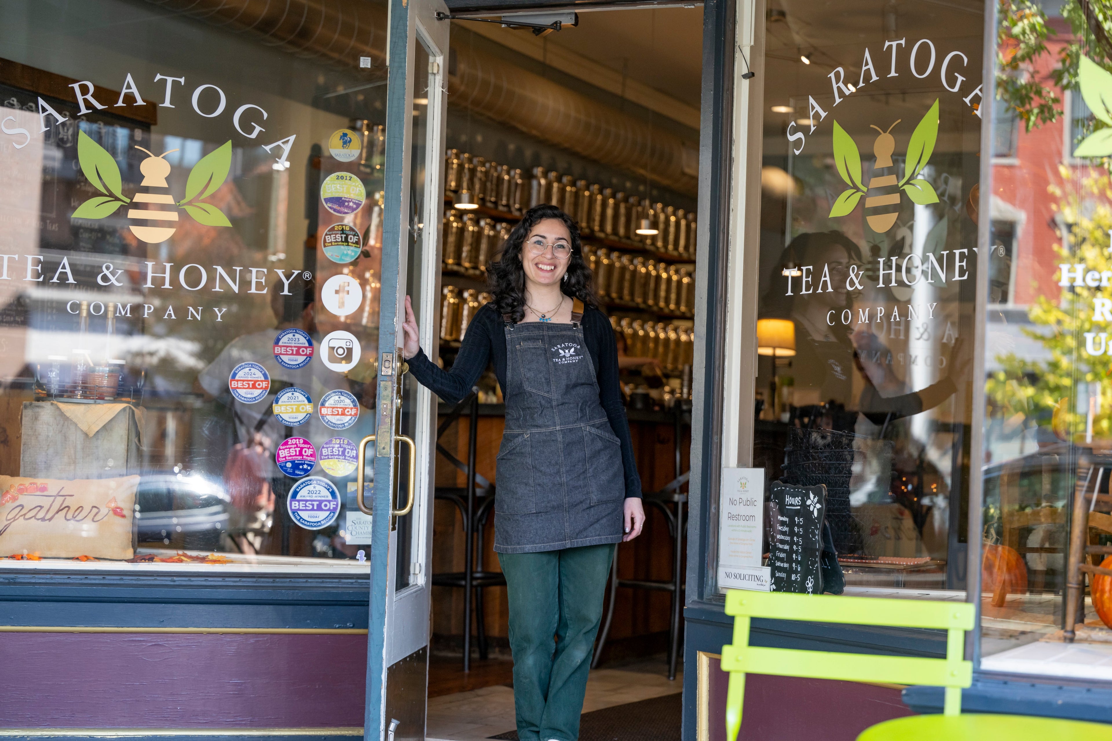 Woman with black hair in a denim Saratoga Tea & Honey Co. apron holding open the door to Saratoga Tea & Honey Co.