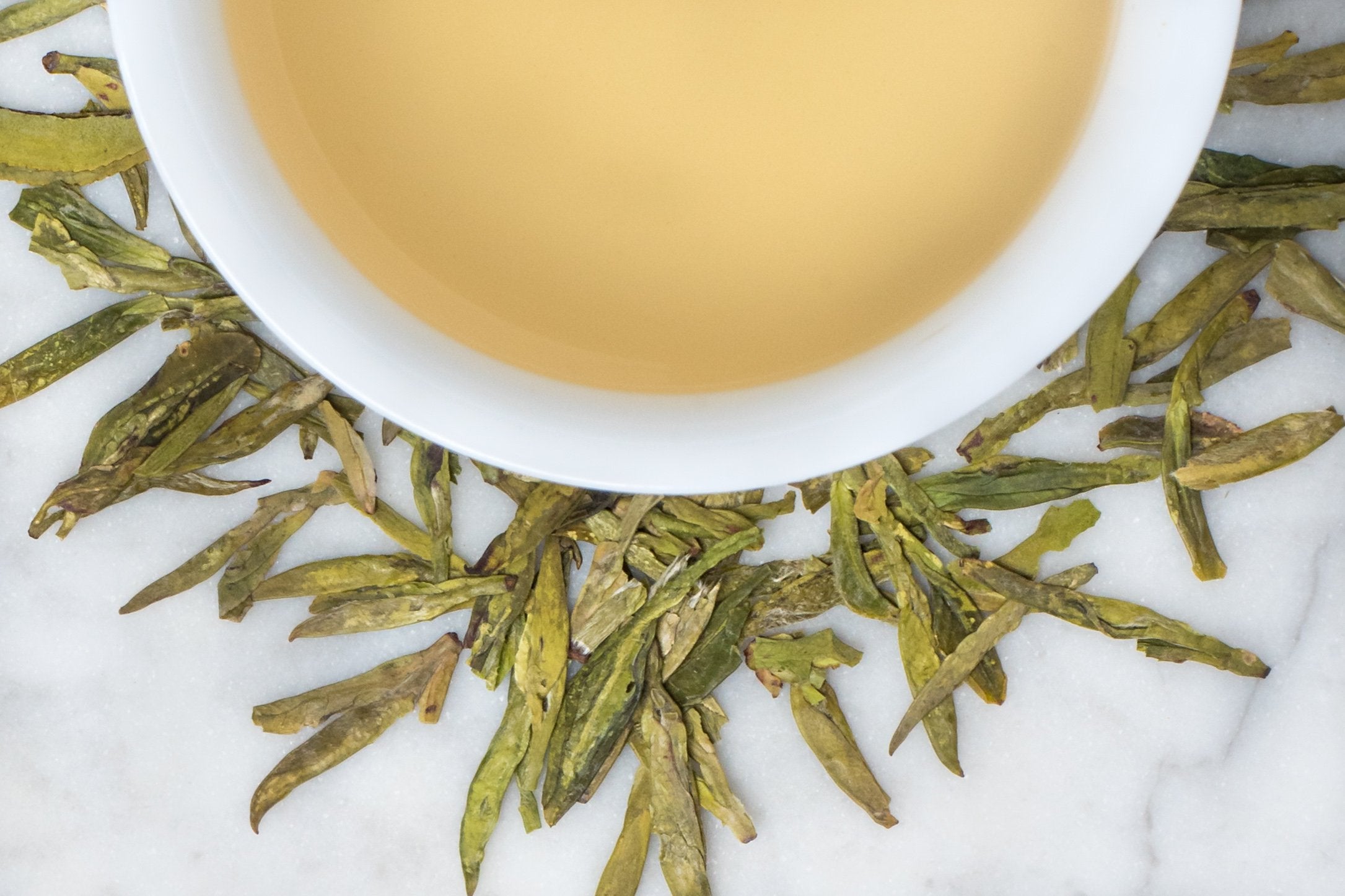 Loose leaf Long Jing Dagonwell green tea surrounding a white cup with brewed green tea