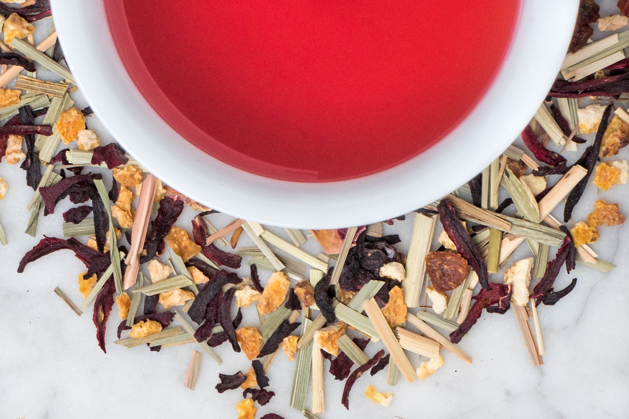 loose leaf hibiscus and citrus herbal tisane surrounding a white cup with brewed tea